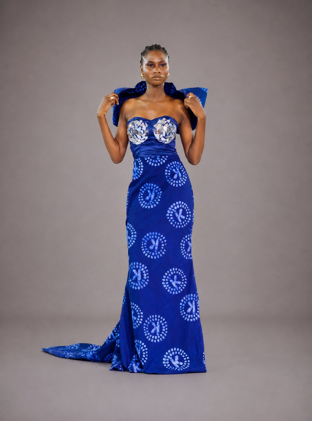 Mannequin portant une robe haute couture IK Tissu sirène longue en wax indigo aux médaillons circulaires blancs. Bustier en V brodé de paillettes miroir, drapé d'épaules sculptural en volume. Pose frontale, mains aux côtés, élégance souveraine.