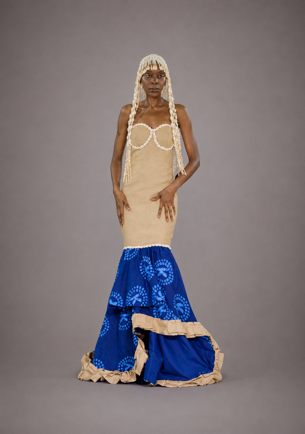 Mannequin portant une robe haute couture IK Tissu, coiffée d'une parure en cauris cream qui descend en cascade jusqu'au buste. Bustier cœur cream à finitions perlées, jupe sirène en wax indigo aux motifs circulaires teints à la main blancs, ourlet contrasté en plissé cream. Silhouette frontale, allure cérémonielle.
