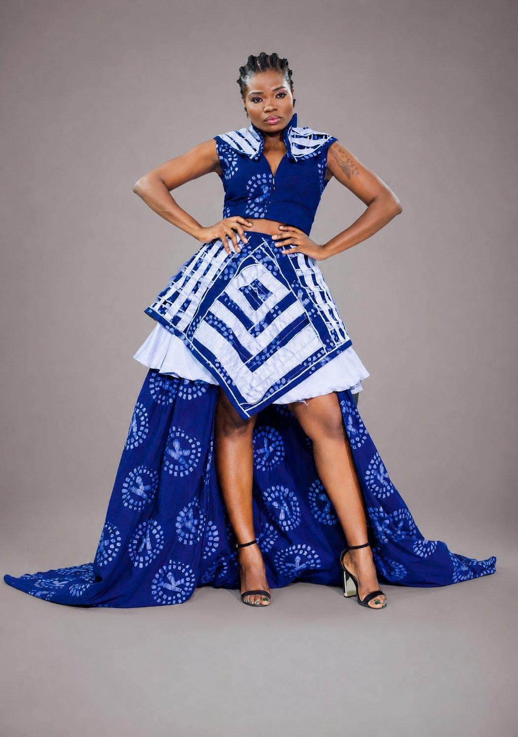 Mannequin portant une robe haute couture IK Tissu de coupe asymétrique : bustier en V profond en wax indigo et blanc, jupe courte cream plissée devant, longue traîne indigo ornée de médaillons circulaires blancs et d'un panneau central à motif géométrique en losange. Pose mains sur les hanches, regard direct, présence affirmée.